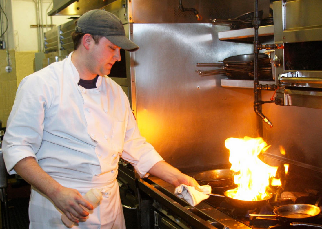 Chef Spencer in front of a range with two pans and a large flame. 